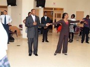 Formally dressed guests at an indoor community gathering, standing in conversational groups in a plainly furnished hall.