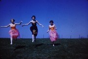 Three young girls in ballet poses against a twilight sky: two in pink tutus and pointe shoes flank a girl in a dark leotard, all with arms raised and extended.