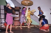 Six female students in school gymnasium perform singkil, stepping between clapped bamboo poles held by two dancers on either side.