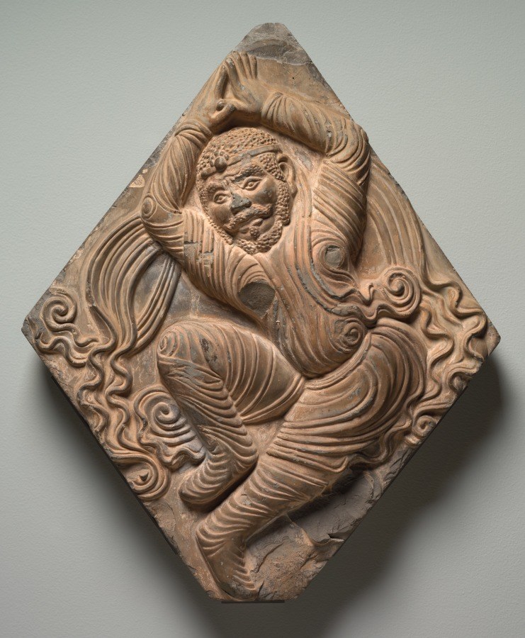Relief carving of a bearded male dancer with raised arm, surrounded by swirling acanthus ornament, set in a diamond-shaped terracotta tile.
