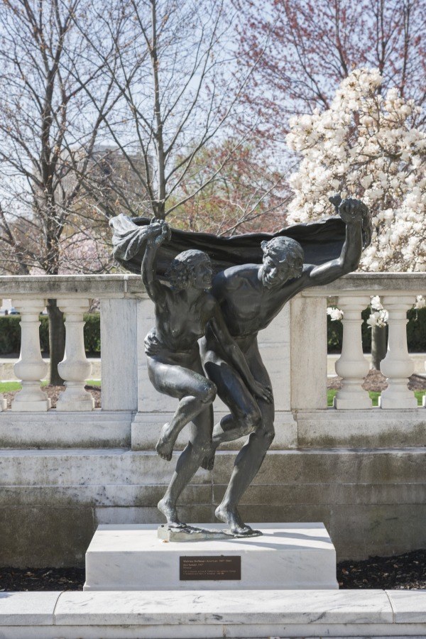 Bronze sculpture of a male figure in mid-leap, body arched backward, one leg extended high, arms sweeping open, holding billowing drapery, set on a stone