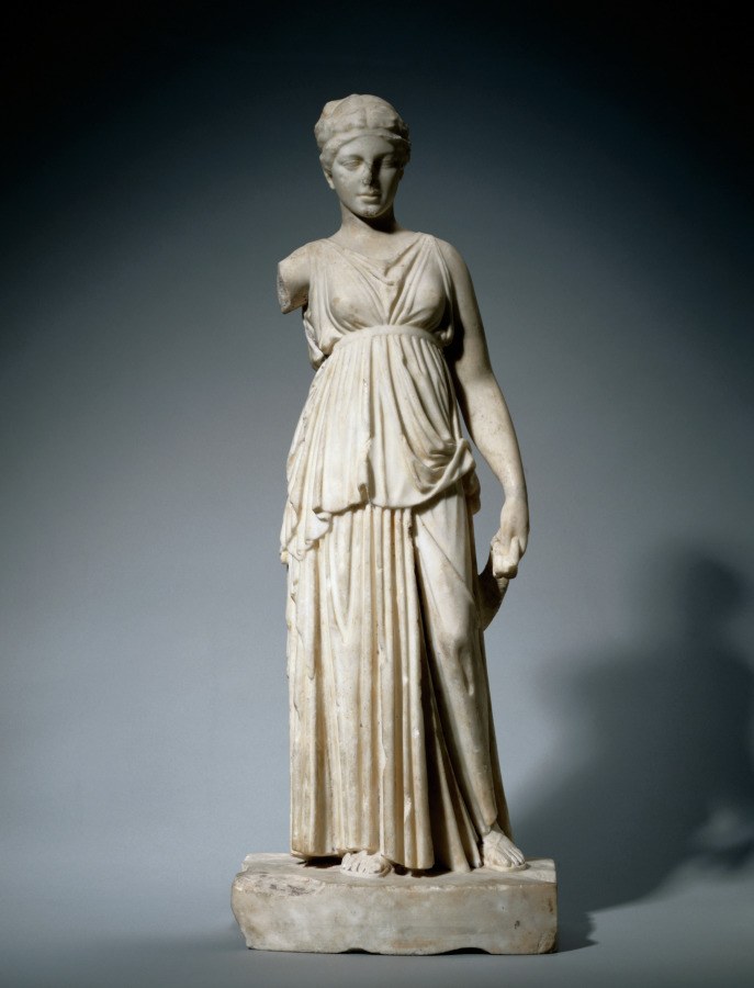 Neoclassical marble sculpture of a draped female figure in contrapposto stance with raised left arm, standing on a cylindrical plinth.