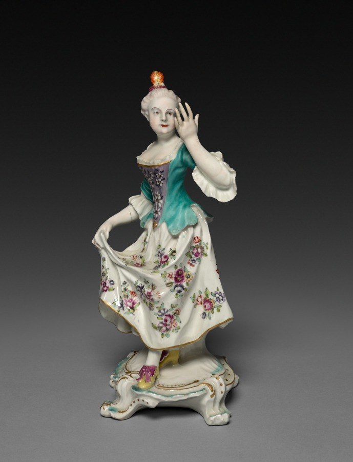 Porcelain figurine of an 18th-century woman in teal bodice and cream silk skirt, hand raised to her head in an elegant pose, standing on a rococo gilt base.