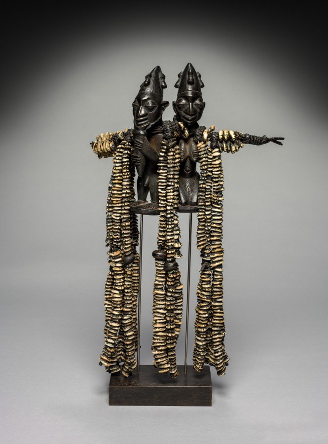 Two mirrored wooden figures with extended arms, wearing stacked brass rings like segmented robes, stand back-to-back on a black base.