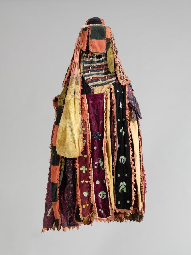 Layered textile masquerade costume with dark cylindrical head covering, draped rust-colored veils, and colorful panels of maroon, gold, and black fabric