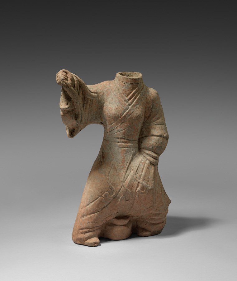 Terracotta figure of a kneeling dancer in a pleated robe, left arm raised with hand near face, head turned to the side.