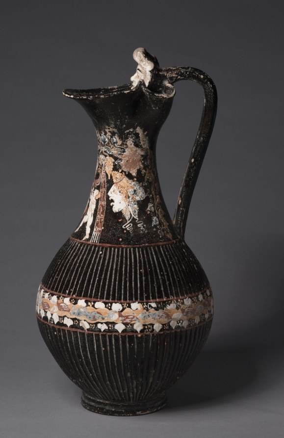Black ceramic Greek wine jug with a flared mouth and high handle, decorated with theatrical masks alternating with dancing figures rendered in golden tan glaze.