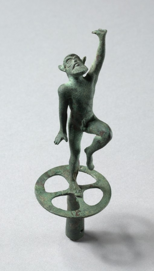 Green patina bronze satyr figurine leaping on one leg with arm raised, mounted on a circular platform with apertures, viewed against white background.