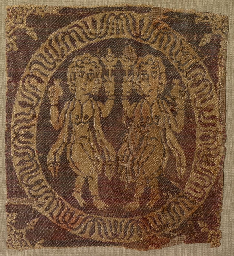 Fragmentary woven textile with two classical female figures in gold thread on reddish-brown ground, bordered by repeating geometric pattern.