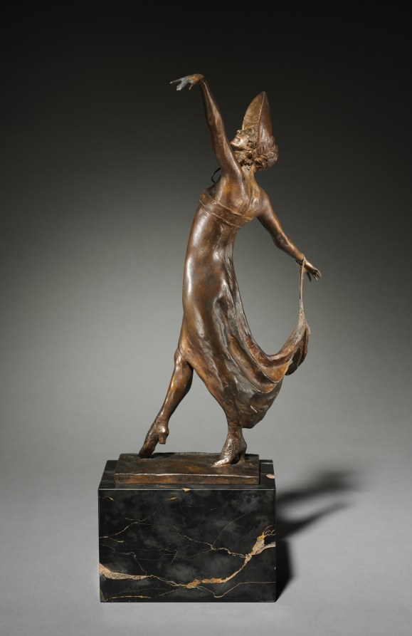 Bronze sculpture of a female dancer in an extreme backward arch on one pointed foot, arms extended, wearing a plumed headdress, on a black marble base.