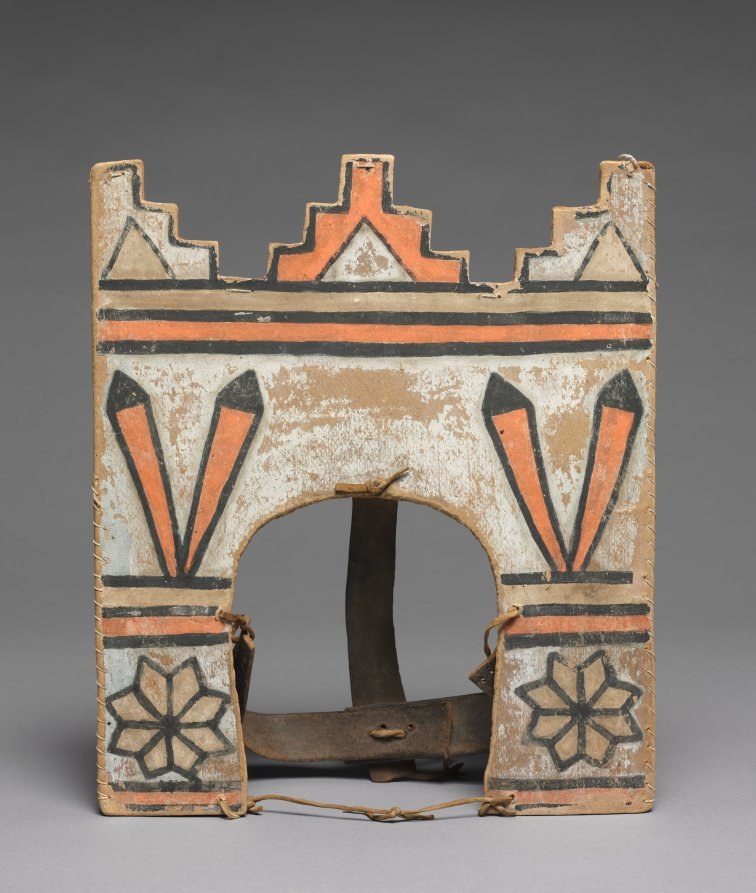 Wooden headdress with geometric painted designs in ochre, black, and orange, featuring a central arch opening and crenellated top edge; iron support visible at