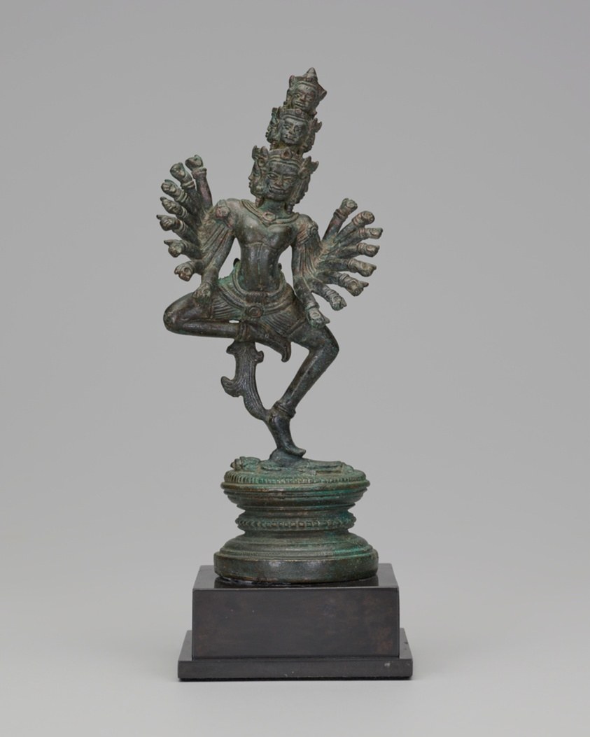 Bronze sculpture of a four-armed dancing deity on a tiered circular base, wearing an ornate tall crown and tilted in contrapposto stance.
