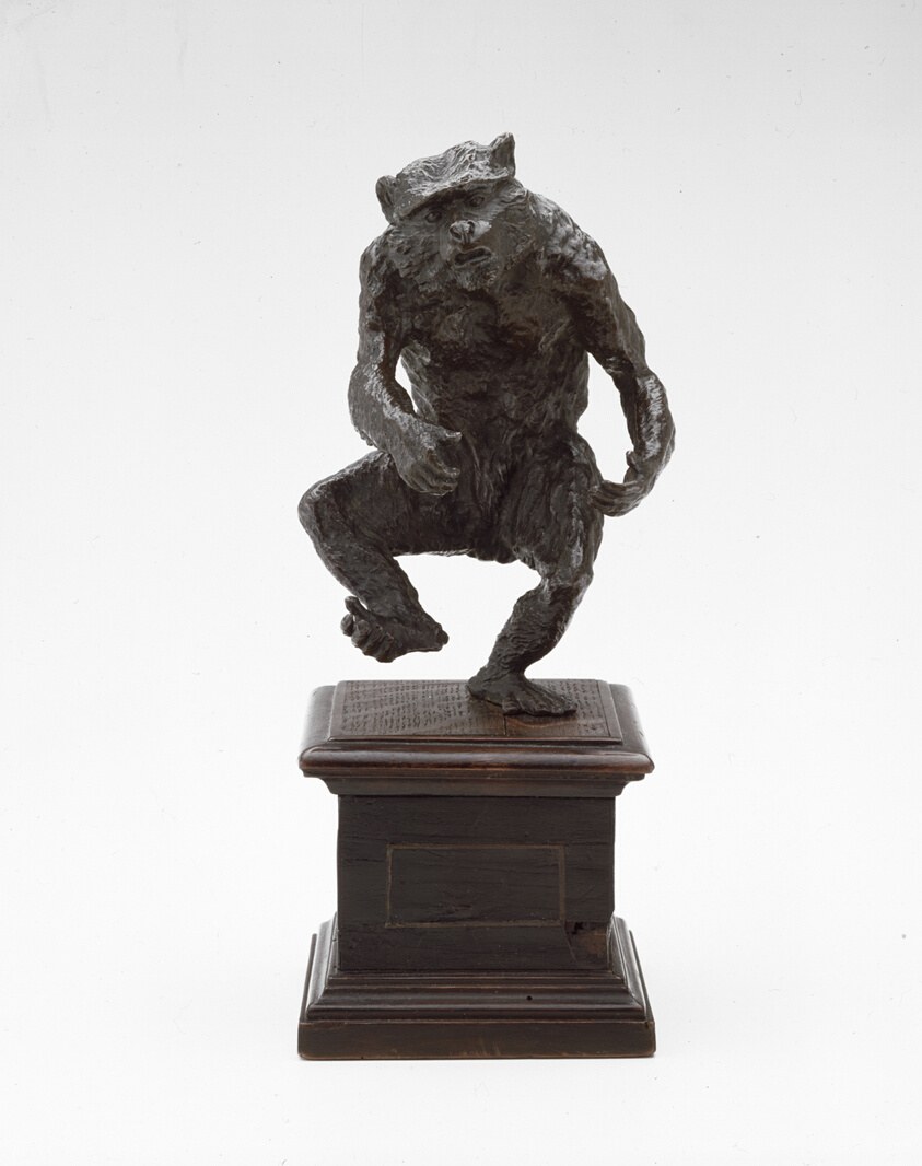 Bronze figurine of a crouching monkey with one arm raised, mounted on a rectangular wooden base with an inscription plaque.