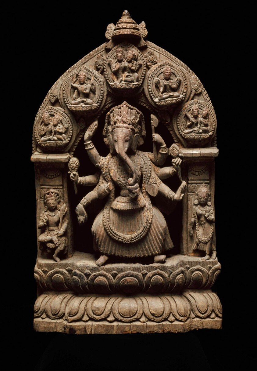 Stone relief of Ganesha dancing with multiple arms raised, flanked by smaller attendant figures in an ornate temple frame.