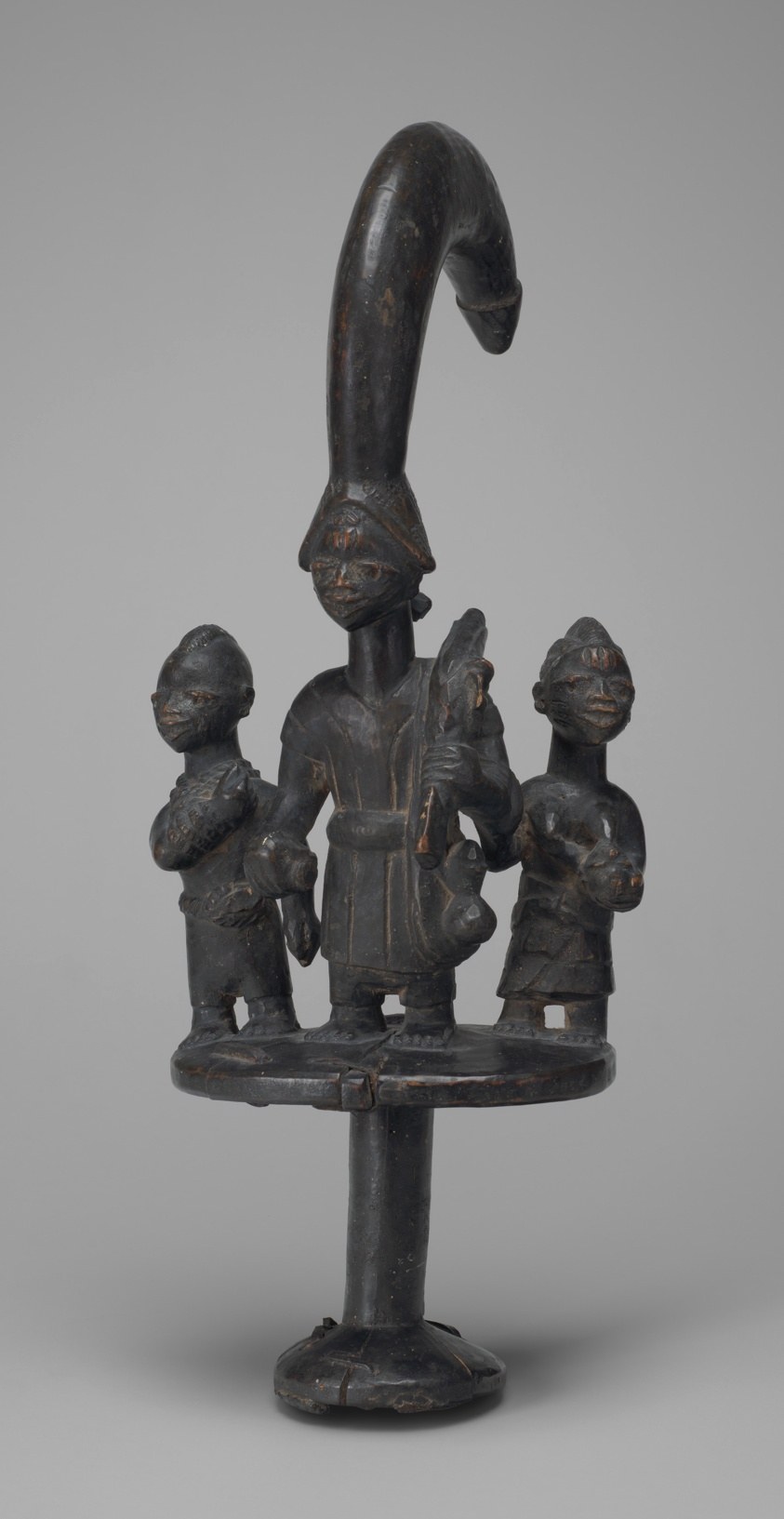 Dark bronze staff with curved finial above a platform holding three small figures: a tall central figure holding a staff, flanked by two smaller attendants.