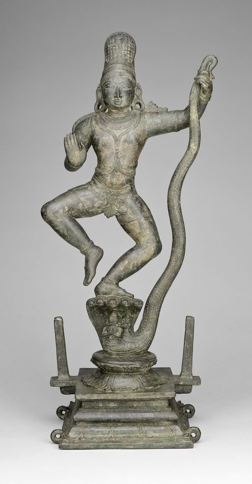 Bronze sculpture: a divine figure in a high-stepping dance pose on a serpent's head, wearing a tall crown and ornate jewelry, arms outstretched.