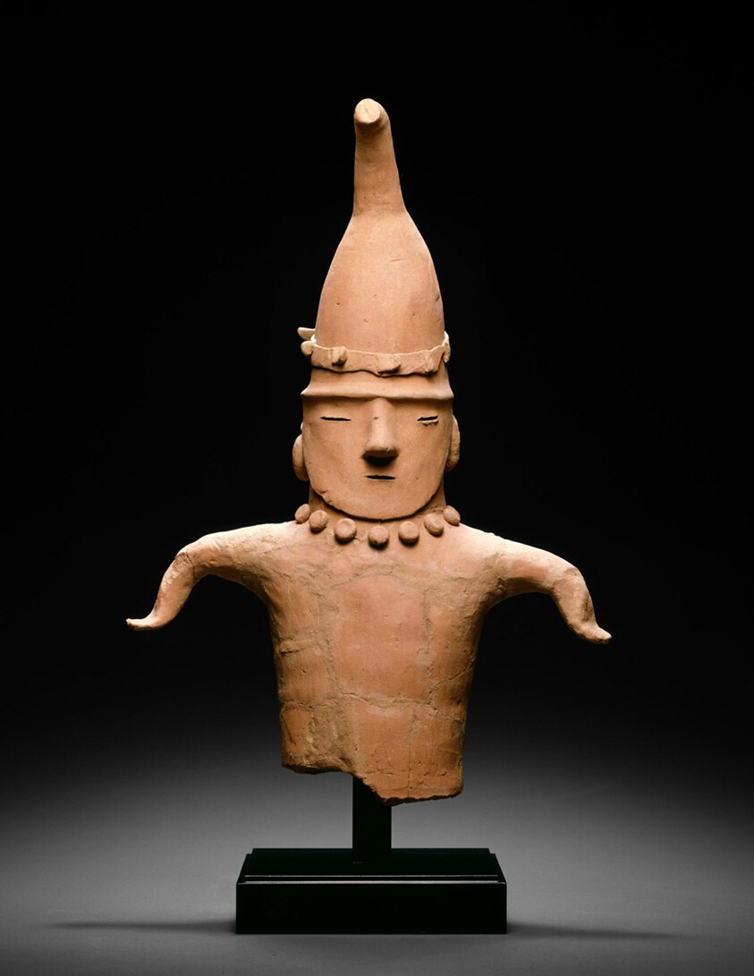 Terracotta sculpture of a standing figure with raised arms, small head, geometric torso, pectoral collar, and tall conical headdress.