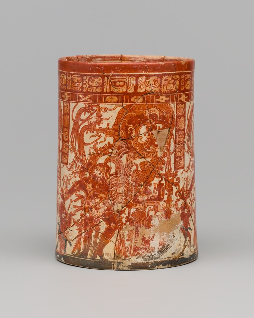 Red and cream Maya ceramic cylinder with figures surrounding a central maize god in elaborate headdress and costume.