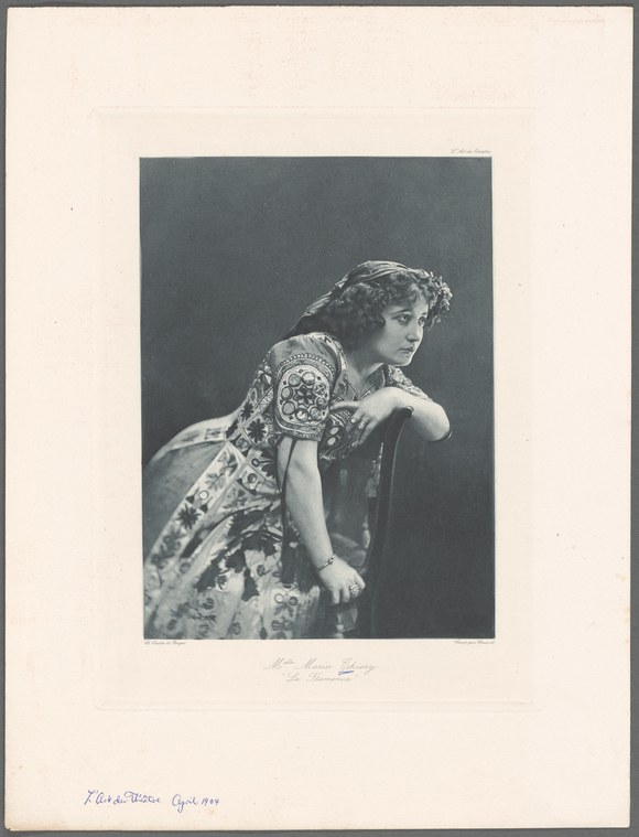 Woman in ornate embroidered dress turns to look over her shoulder in a studio portrait, circa 1890s–1905.