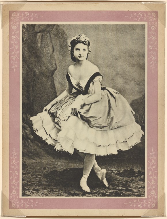 A young woman in a pale tulle ballet skirt and dark corseted bodice stands in a classical ballet pose in a studio setting.