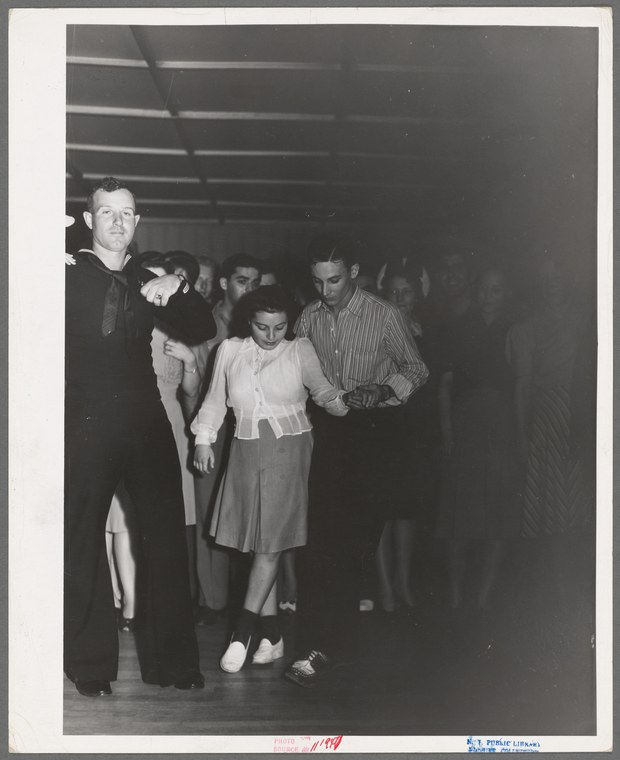 Black-and-white photograph of a man and woman standing together at an indoor dance hall, surrounded by other people, mid-20th century.
