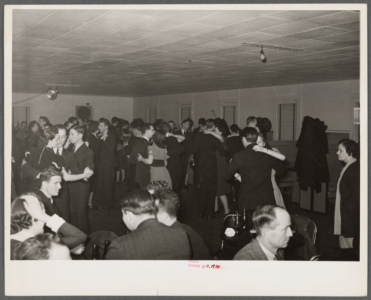 Crowded interior of a public dance hall with dozens of formally dressed couples dancing, photographed from above.