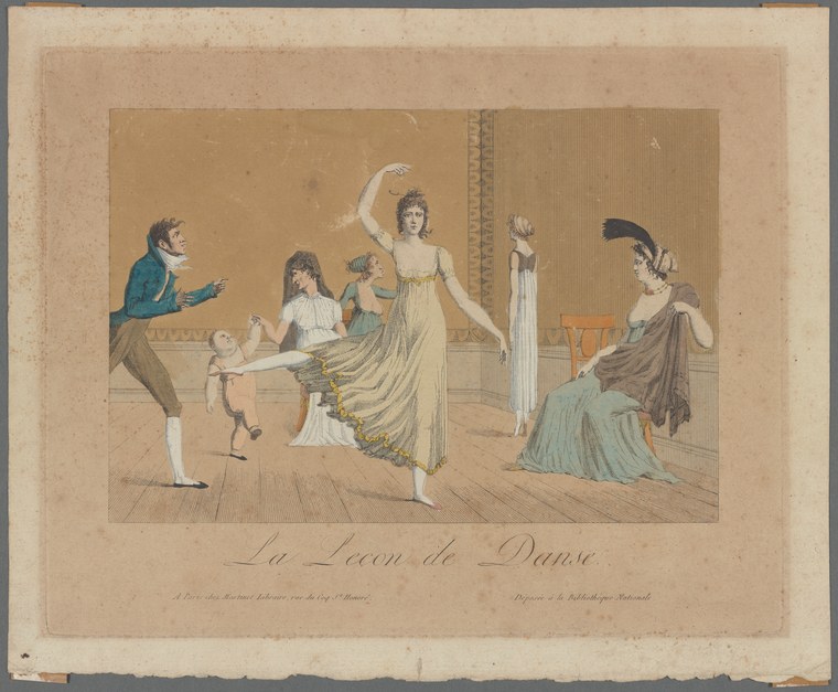 A young male dancer in blue coat lifts his leg high as a female instructor in white raises her arm in correction, with observing students and a chaperone in a
