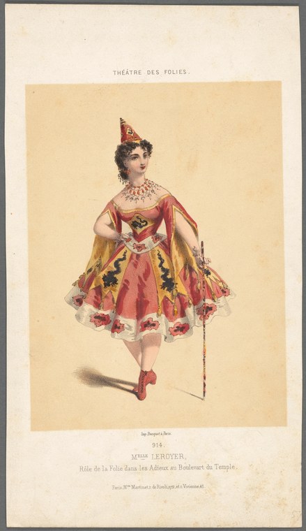 A woman in an ornate red and gold costume with a pointed headdress holds a decorated cane, posed frontally against a pale yellow background.