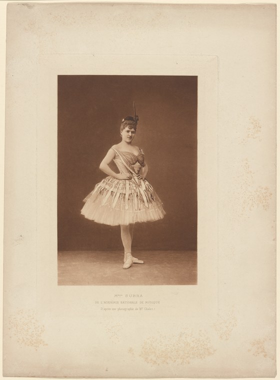 A young woman in 19th-century ballet costume—white tulle skirt and fitted bodice—stands in classical pose against a brown studio backdrop.