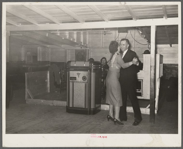 A couple in a closed ballroom hold dances indoors in a plain, concrete-floored dance hall with overhead beams and support columns.