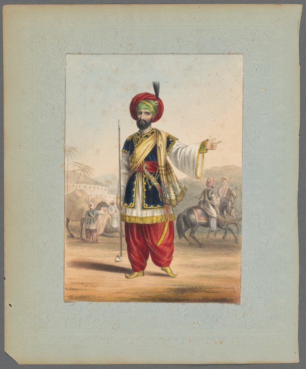 A man in red turban, yellow tunic, and red trousers stands holding a staff with his right arm extended in formal gesture, set against a sparse landscape.
