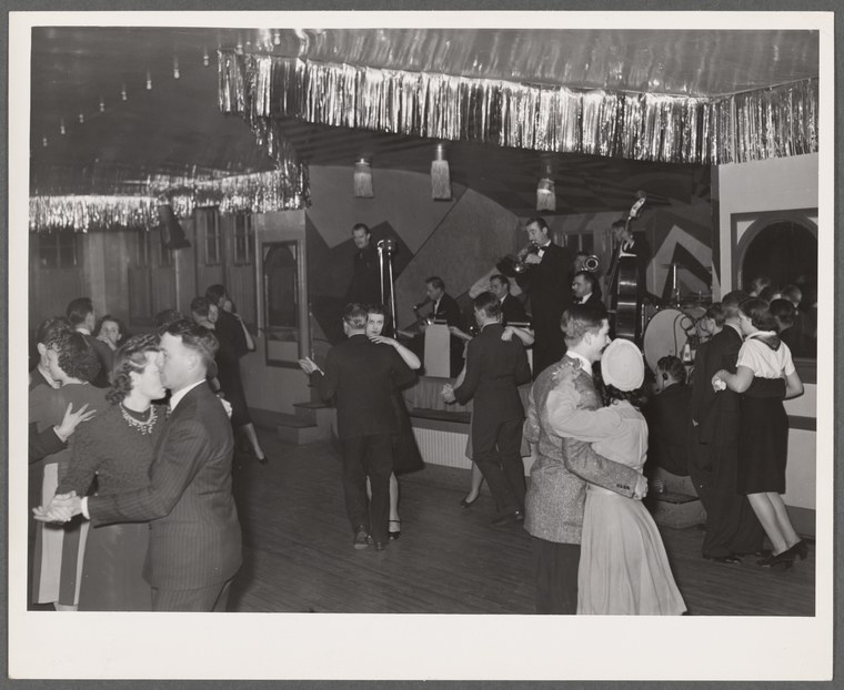 Black-and-white photograph of couples dancing in a crowded, decorated dance hall with overhead streamers.