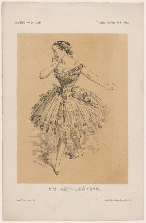 A young ballet dancer in pointe shoes and tulle skirt stands in arabesque pose, one arm raised to her head, against a tan background.
