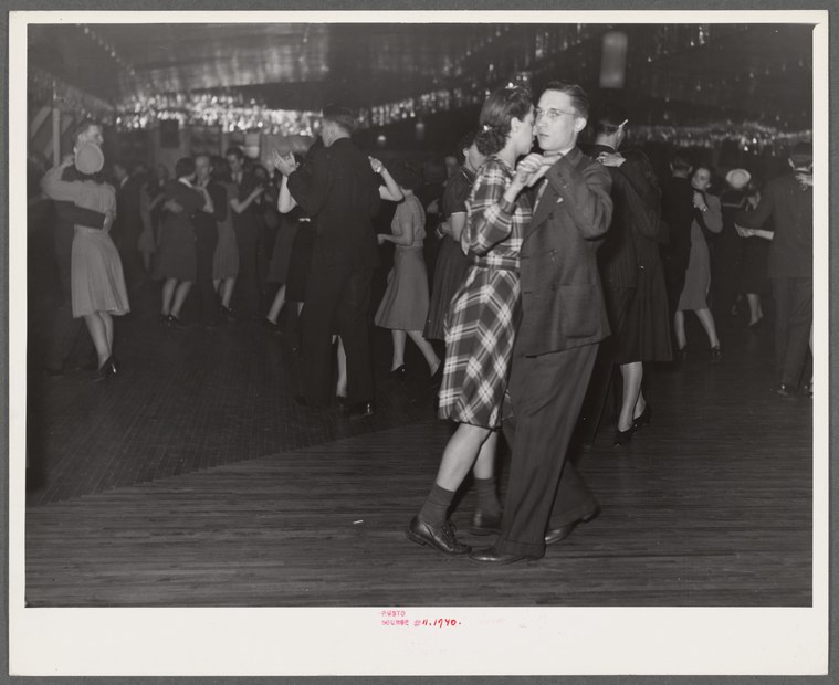 Crowded dance hall with couples dancing in closed ballroom position; foreground couple in plaid dress and suit moves across wooden floor under bright lights.