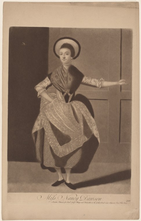 Woman in 18th-century dress with ruffled sleeves and patterned skirt stands in theatrical pose with one arm extended, wearing a wide-brimmed hat.