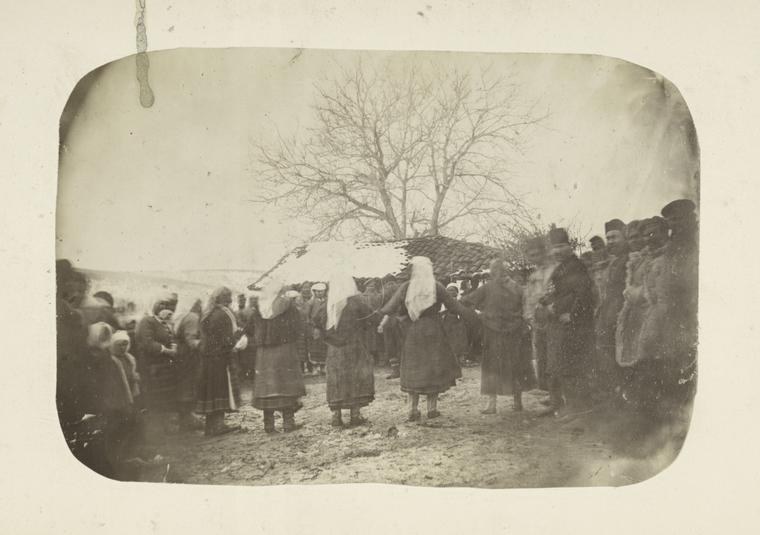 A large group of people in dark winter coats stand outdoors in a loose formation with a bare tree behind them.