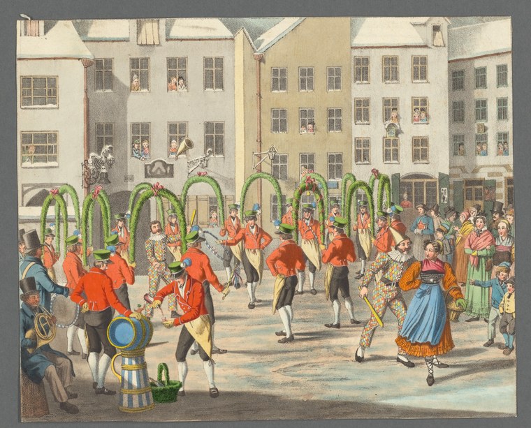 Courtyard surrounded by buildings with dozens of people in bright costumes jumping and moving around seven green woven arch structures.