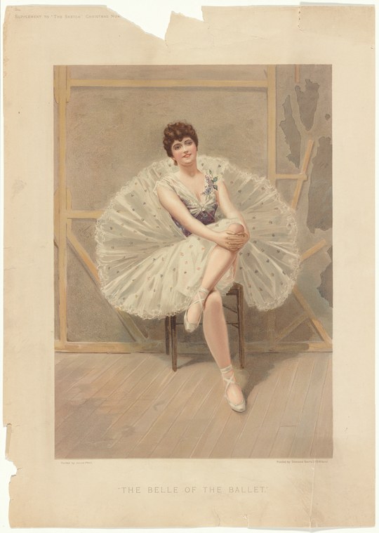 Young female ballet dancer in white tutu seated on wooden chair, wearing pointe shoes, looking toward viewer with composed expression.