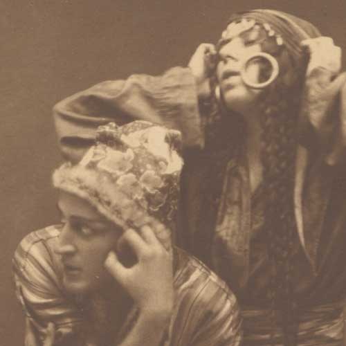 Sepia-toned photograph of two dancers in a theatrical pose: one reclines with hand to face while the other bends forward in an embrace.