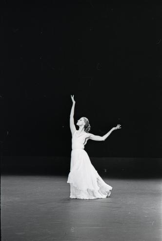 A solo female dancer in a white flowing dress stands alone on a dark stage, head tilted back, arms extended open, in an expressive pose.