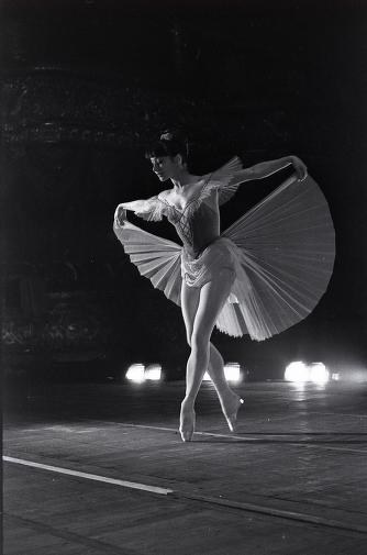 Ekaterina Maximova in pointe shoes and classical tutu performs an extended arabesque against a dark stage, lit by spotlights.