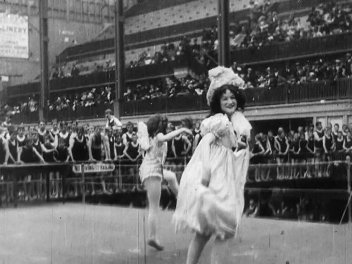 Black-and-white stage photograph of a female dancer in a white Regency-era gown with feathered headdress, arms extended, performing before an audience.
