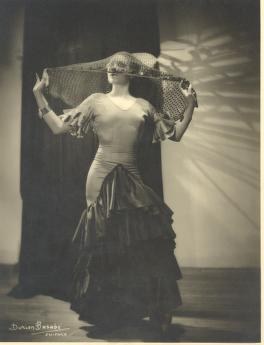 Studio photograph of Katherine Dunham in 1920s evening dress, holding a large fan overhead, posed against a dark interior.