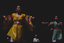 Classical Indian dancers in vibrant traditional costumes perform at a Chicago plaza festival, striking formal poses with lifted frames and extended arms.