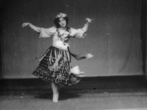 Female dancer in white blouse and patterned dark skirt, arms extended, mid-turn on stage, 1898.