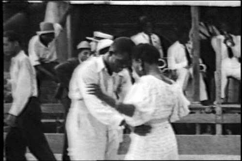 Black-and-white photograph of Jamaican couples dancing in close embrace outdoors, surrounded by standing onlookers in light clothing.