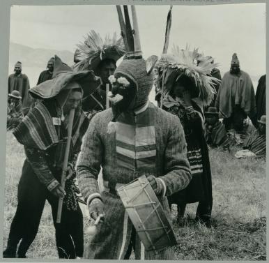 Group of Native Americans in traditional regalia including feathered headdresses and woven blankets stand in a grassland gathering.