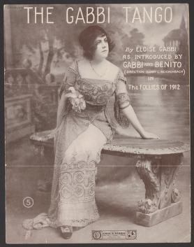 Woman in white lace dress seated at a small table, wearing long gloves, posed for a 1912 theatrical portrait.