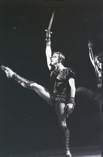 A male ballet dancer in mid-leap against a dark background, right arm raised holding a sword, left leg extended high.
