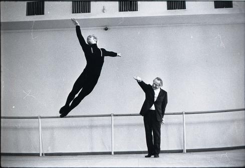 Two dancers in a bare studio: one leaps horizontally with extended limbs while the other stands watching, arm raised.